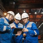 Grupo de engenheiros discute um projeto em um tablet dentro de uma fábrica.